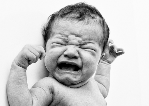 A black and white image of a newborn baby crying with a scrunched-up face and arms in the air.