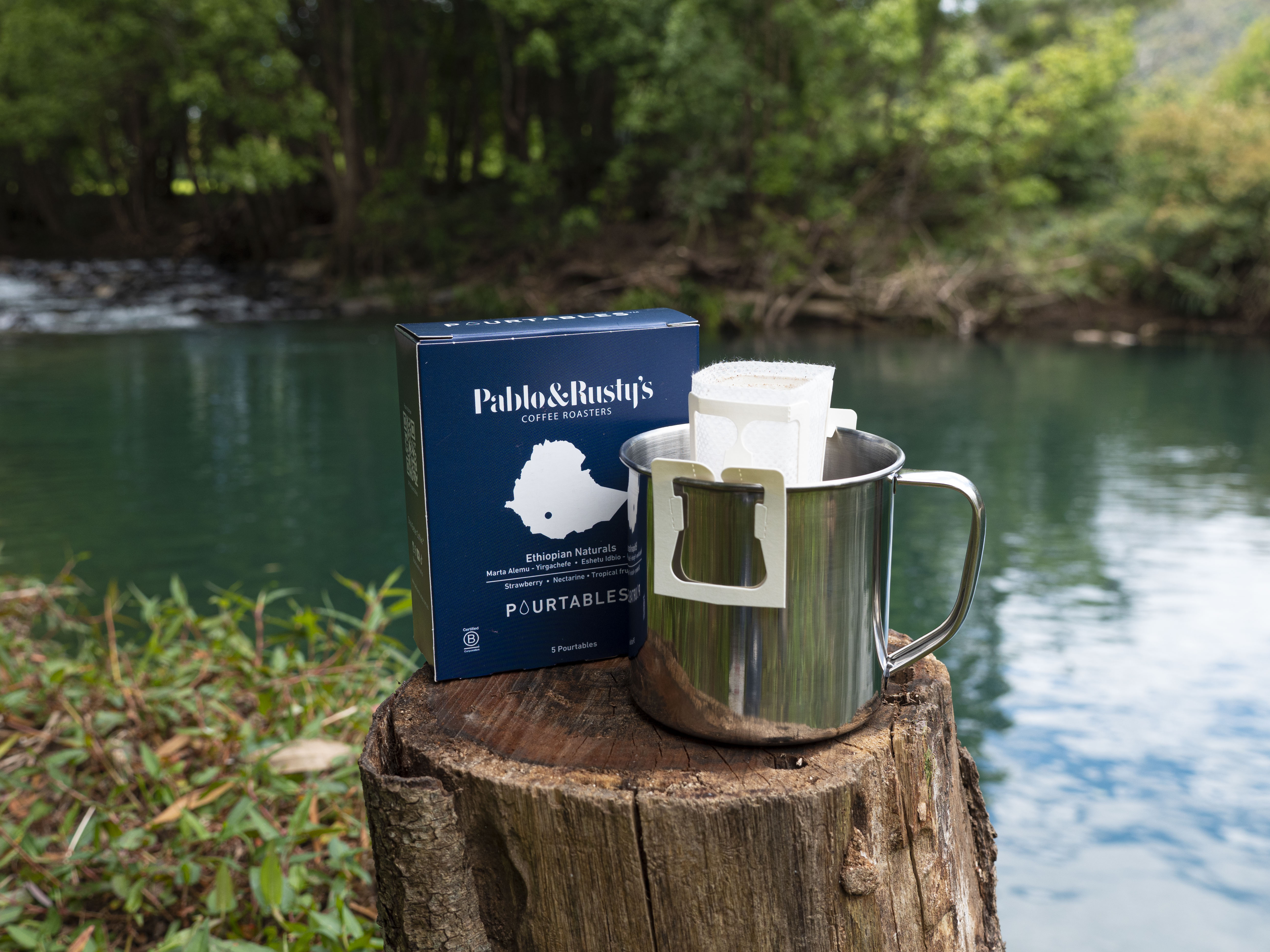 Pack of P&R Ethiopian Natural Pourtables resting on a log next to a cup, with a blue lake behind. 