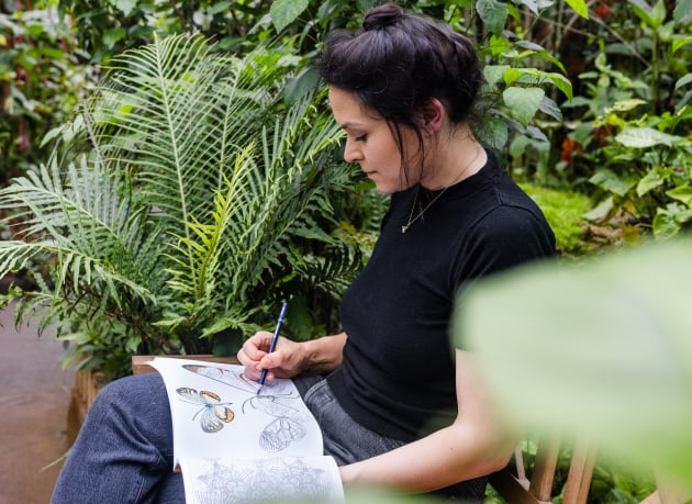 A woman sits on a bench with an adult coloring book surrounded by lush greenery. 