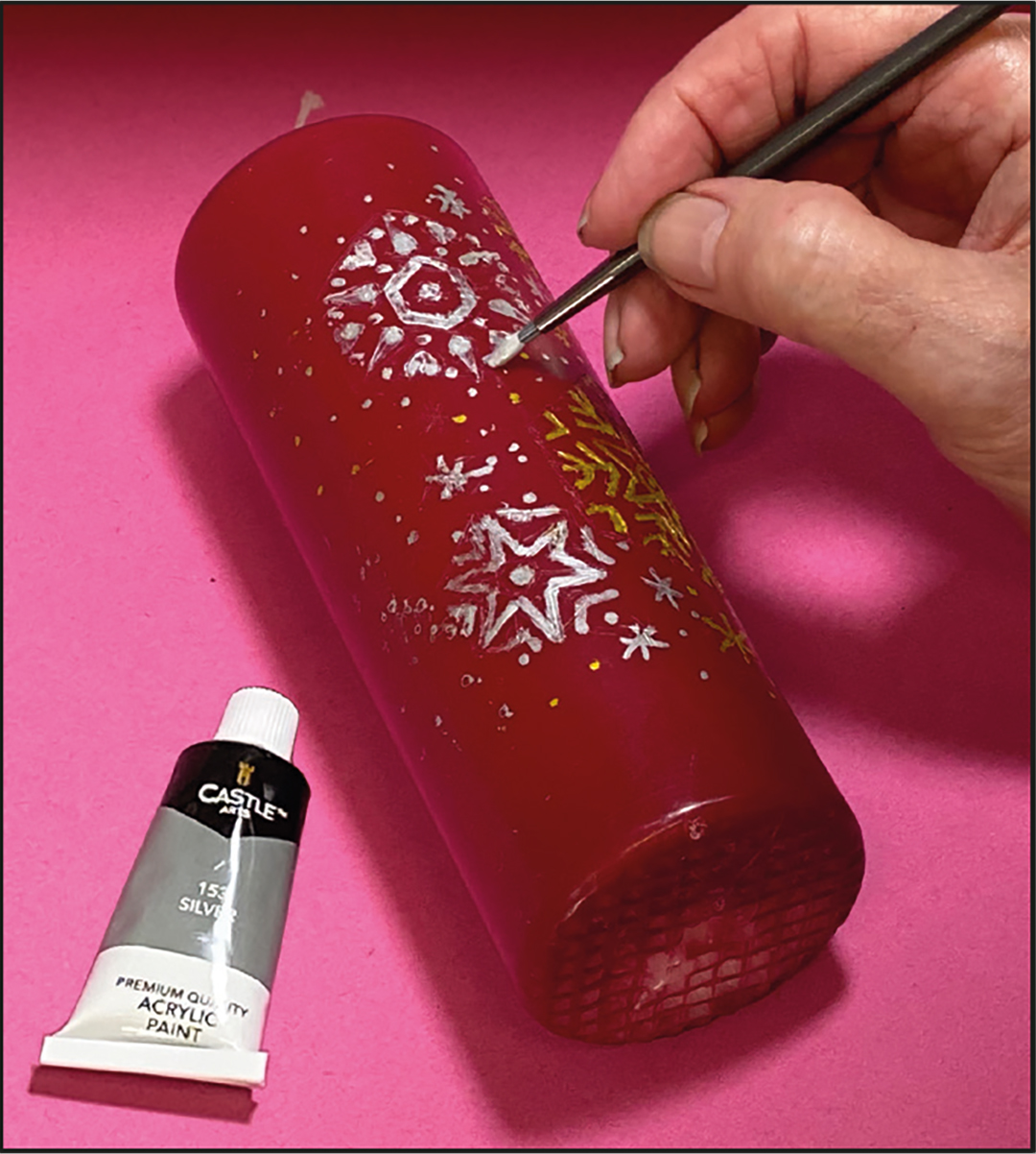 An artist painting over the pencil indentations on a Christmas candle with acrylic paint.