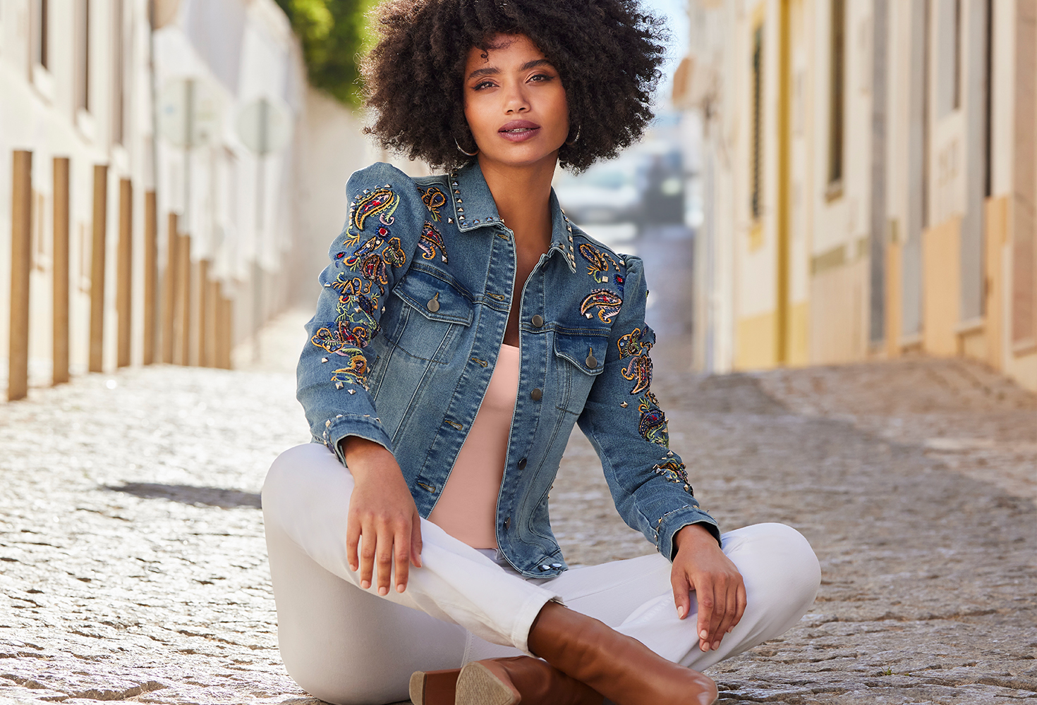 model wearing an embroidered and embellished puff-sleeve denim jacket, pink top, silver hoops, white jeans, and brown heeled leather booties.