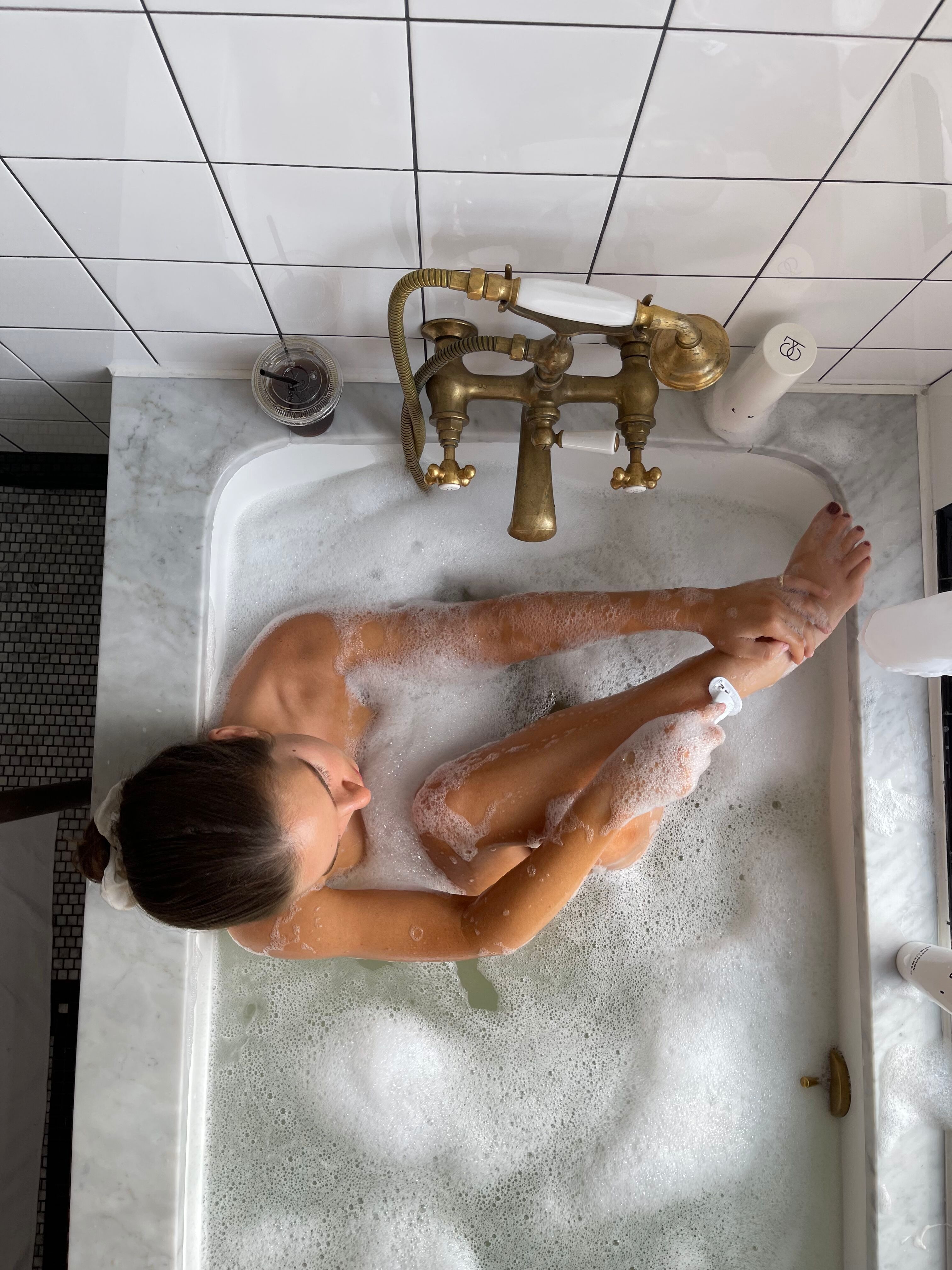 Woman shaving her leg while relaxing in a bubble bath.