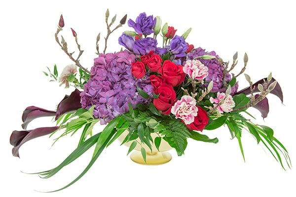 A Bespoke style centerpiece features red roses, purple hydrangeas, pink and white striped carnations, chocolate calla lilies, blossoming spring branches, and an assortment of foliage in a gold compote.