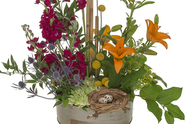 This beautiful floral design mixes orange lilies, craspedia, red blooms, eryngium, salal, Italian ruscus, and features a petite bird's nest with tiny eggs.