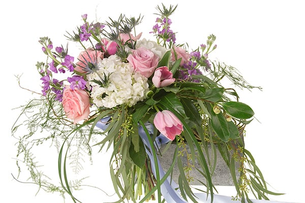 A beautiful Bespoke style hand tied bouquet mixes pink roses, white hydrangea, pink tulips, blue eryngium, purple freesia, seeded eucalyptus, and other foliages, and is tied off with a pale blue ribbon.