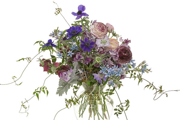 A spectacular Bespoke style spring garden bouquet in an elegant glass vase mixes sweet peas, viburnum berries, tweedia, scabiosa, hellebores, anemones, ranunculus, dusty miller, and passion vine.