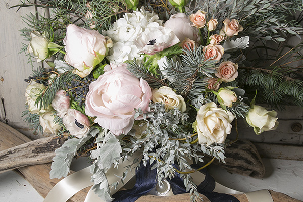 A gorgeous hand tied bouquet features pale pink peonies, cream roses, peach spray roses, white hydrangea, dusty miller, evergreens, and other foliages, all tied off with an ivory satin ribbon.