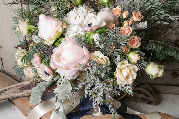 A gorgeous hand tied bouquet features pale pink peonies, cream roses, peach spray roses, white hydrangea, dusty miller, evergreens, and other foliages, all tied off with an ivory satin ribbon.