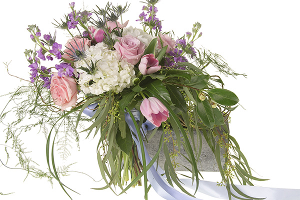 A beautiful Bespoke style hand tied bouquet mixes pink roses, white hydrangea, pink tulips, blue eryngium, purple freesia, seeded eucalyptus, and other foliages, and is tied off with a pale blue ribbon.