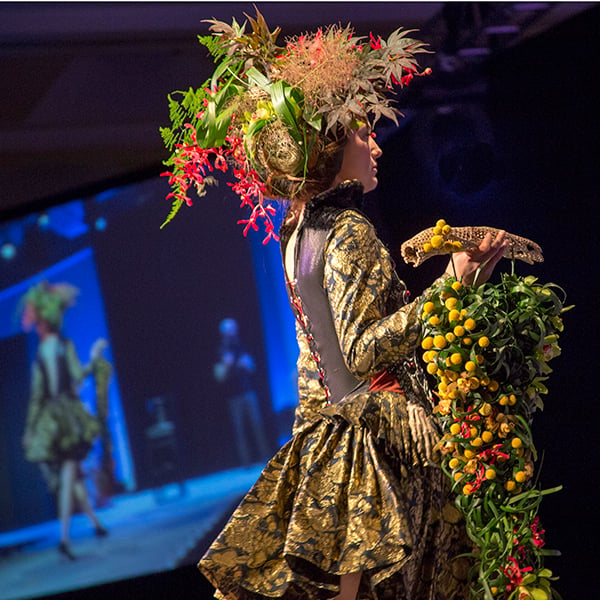 At a fashion show, a model wears a stylish headpiece composed of flowers and foliage, and carries a handbag covered in floral materials.