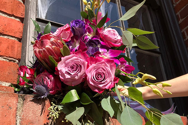 A beautiful wedding bouquet in jewel tones features pink roses, purple tulips, coral protea, orchid blossoms, blue eryngium, seeded eucalyptus, and salal.