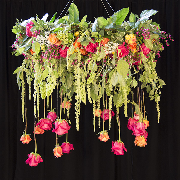 An elegant and opulent floral hanging chandelier combines a variety of blooms in vibrant colors with various foliages intended to create a draping and flowing style. 