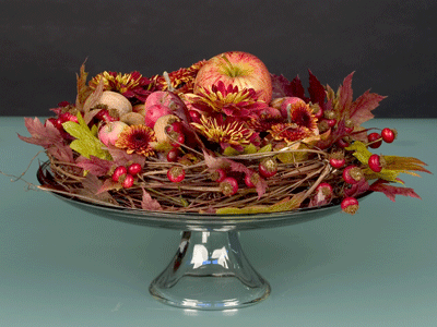 Materials for this design include a grape vine wreath, rose hips, apples, nuts, autumn chrysanthemums, maple leaves, and a pedestal cake plate.