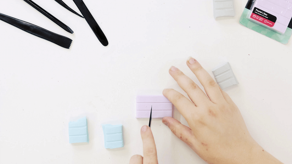 A hand cuts slabs of pastel blue and pastel pink polymer clay in half using a black plastic clay knife.