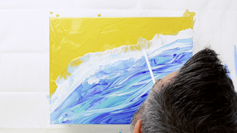 Straw is used to blow the white acrylic pour paint around the canvas to create waves in the beach scene.