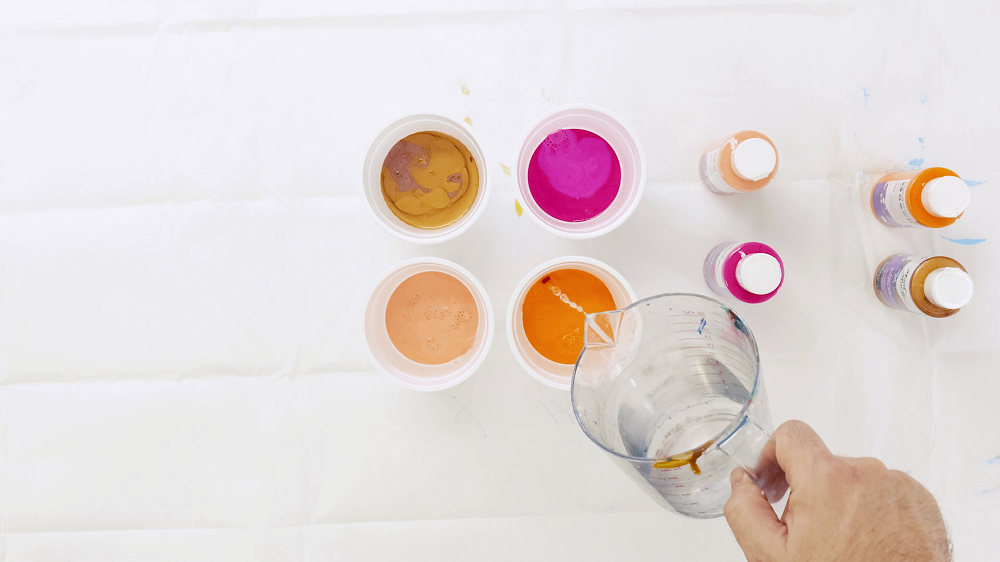 Hand pouring water from a jug into a plastic cup to dilute the pouring paint inside. 