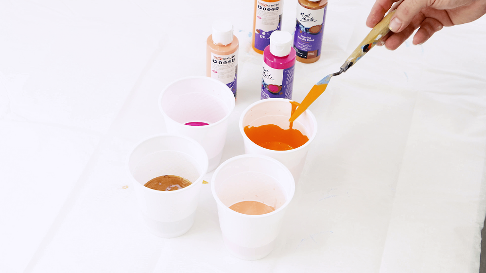 Hand mixes pouring paint with a Mont Marte palette knife, shot shows four plastic cups each filled with different coloured pouring paint.