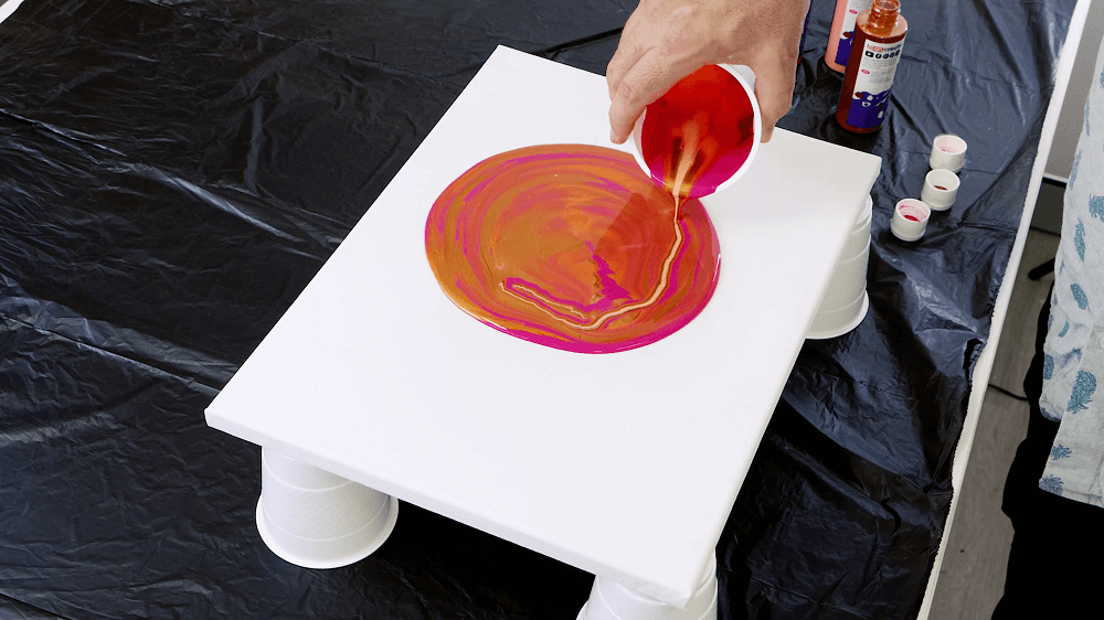 Hand pours a cup full of coral pour paint onto the canvas.