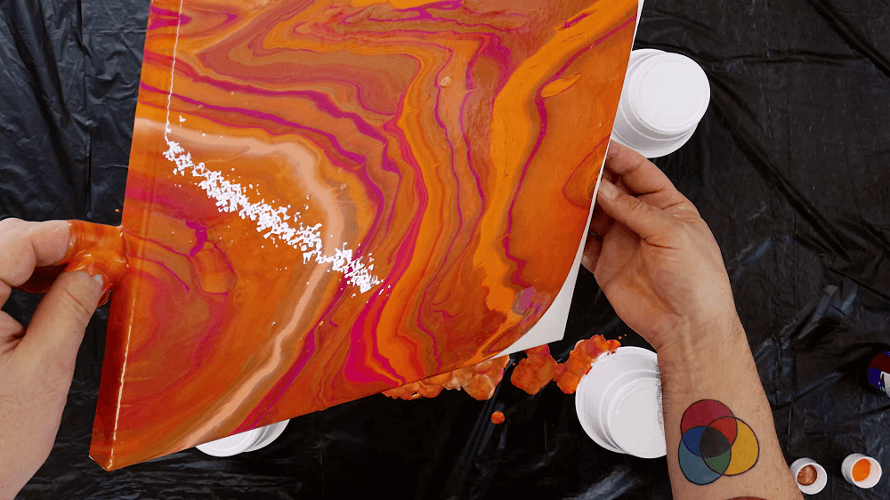 Hand tilts the canvas to spread the coral, orange and pink pour paint to cover the canvas.