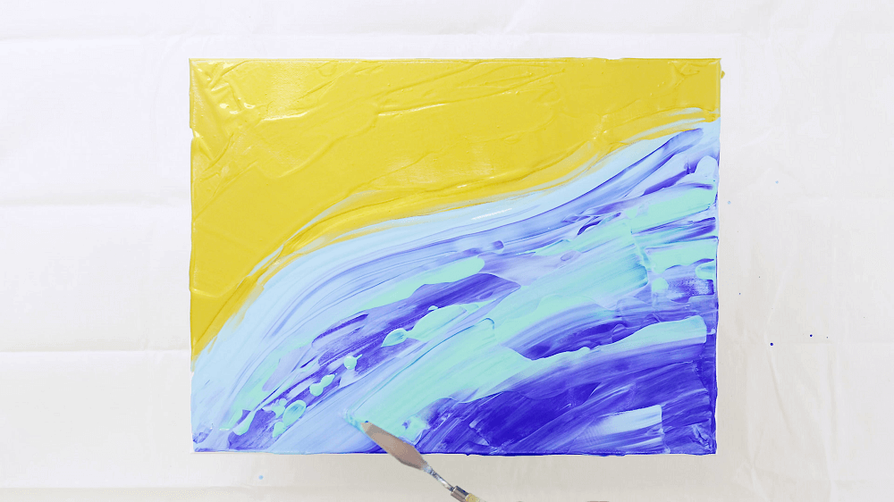 Palette knife blending the yellow and blue acrylic pour paints into a beach scene.