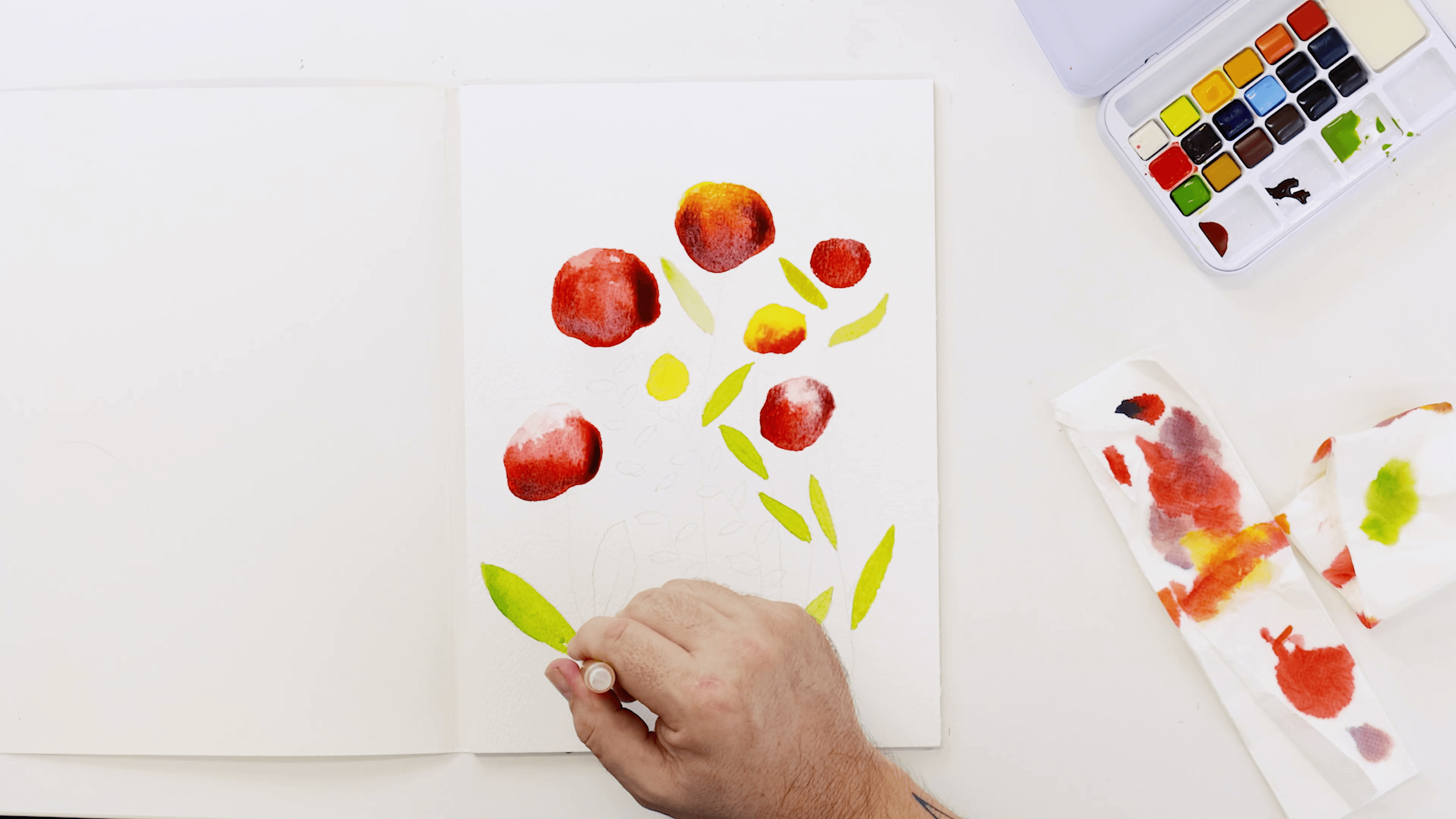 A hand holding a watercolour brush adds Sap Green to darken the leaves.