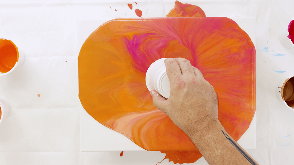 Hand lifts the plastic cup off of the canvas to show paint spilling onto the canvas.