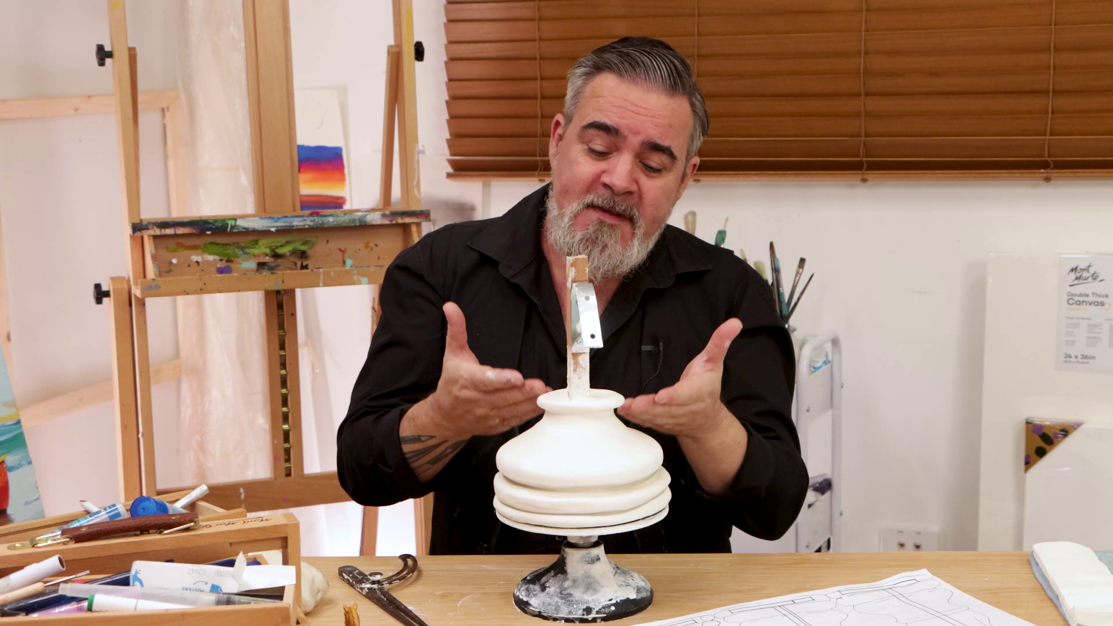 Artist gesturing to clay base of sculpture of pottery wheel