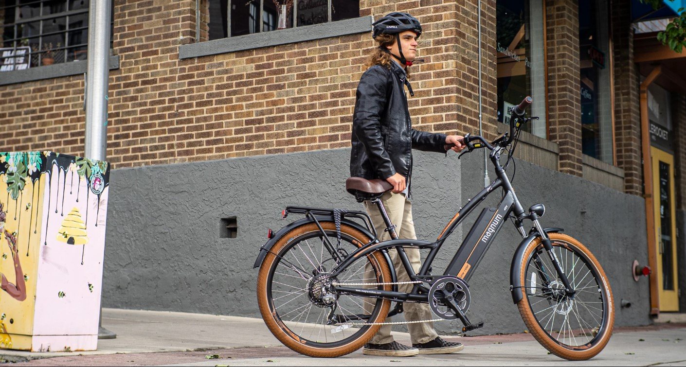 Rider with long hair black jacket khaki pants checkered sneakers wearing helmet and holding up a Magnum Low Rider e-bike