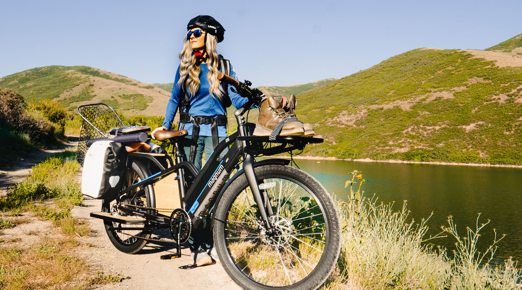 Person with long blonde hair in blue long-sleeve shirt standing next to Magnum Payload cargo e-bike with boots and other gear mounted to e-bike in front of lake and hills