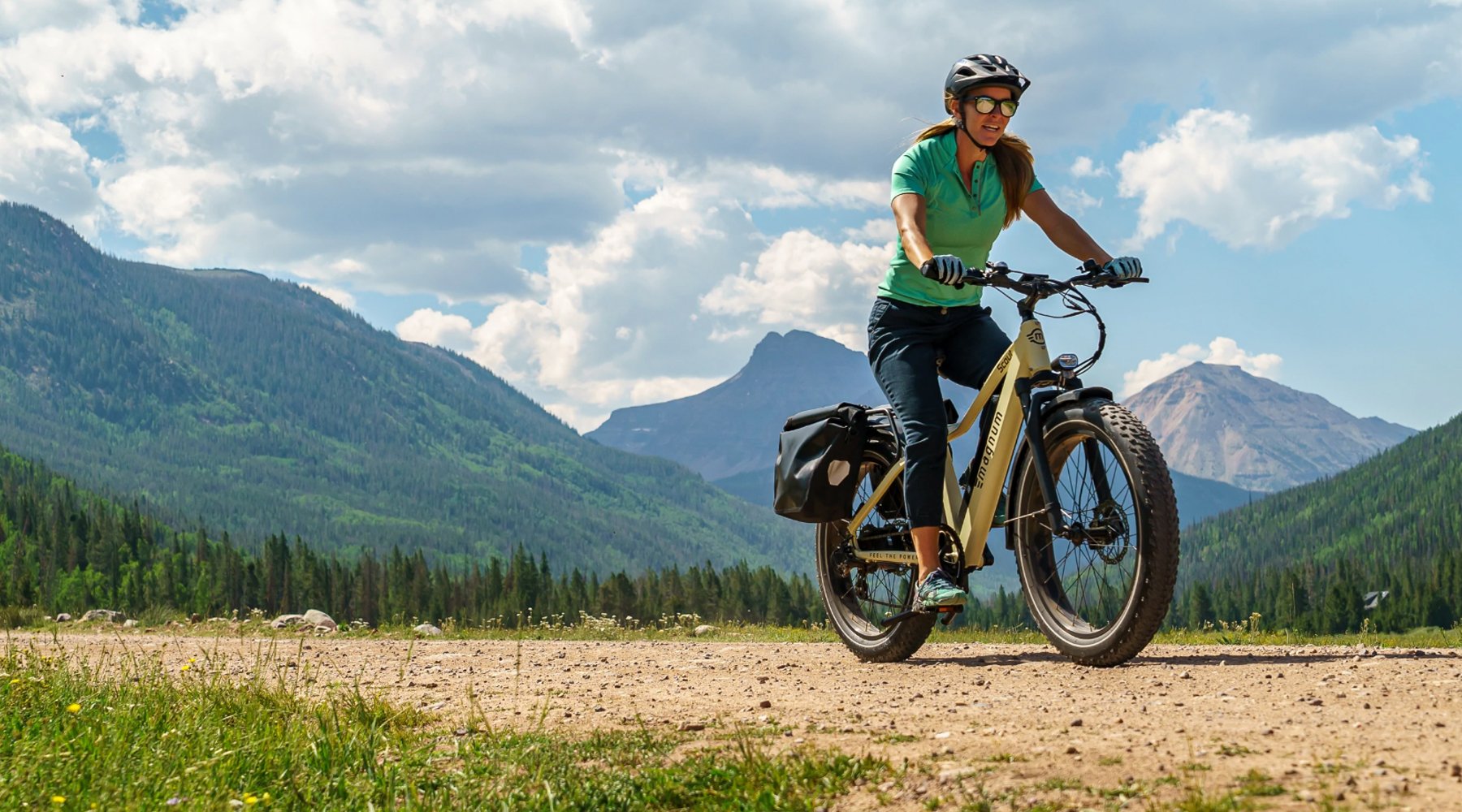 Rider on Magnum Scout electric bike in green polo shirt denim capri pants on dirt trail with mountains blue sky white clouds