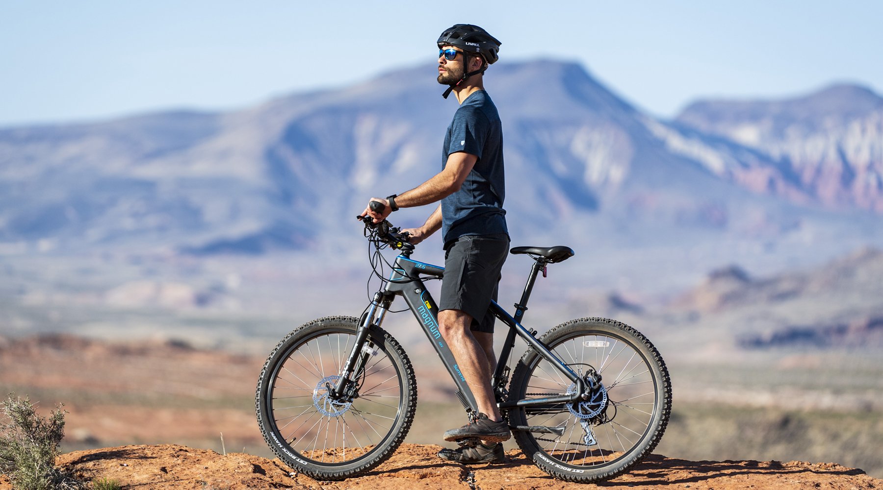 Rider with blue t-shirt black shorts standing over Magnum Summit e-bike looking into the distance desert hills in background