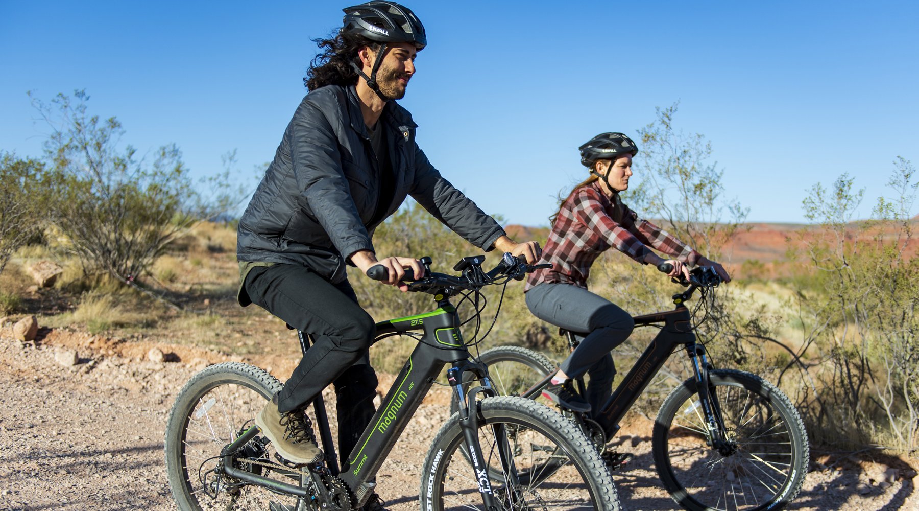 Two riders on Magnum Summit e-bikes on dirt gravel trail with sparse foliage and blue sky