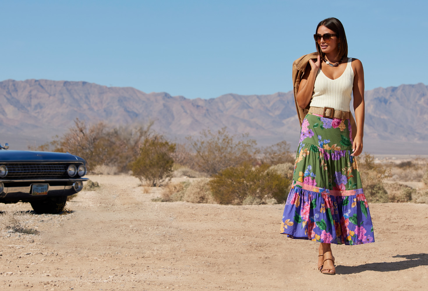 Model wearing an off-white wide ribbed sweater tank top, brown belt, multicolor floral print maxi skirt, tan sandals, and sunglasses with a brown jacket slung over her shoulder.