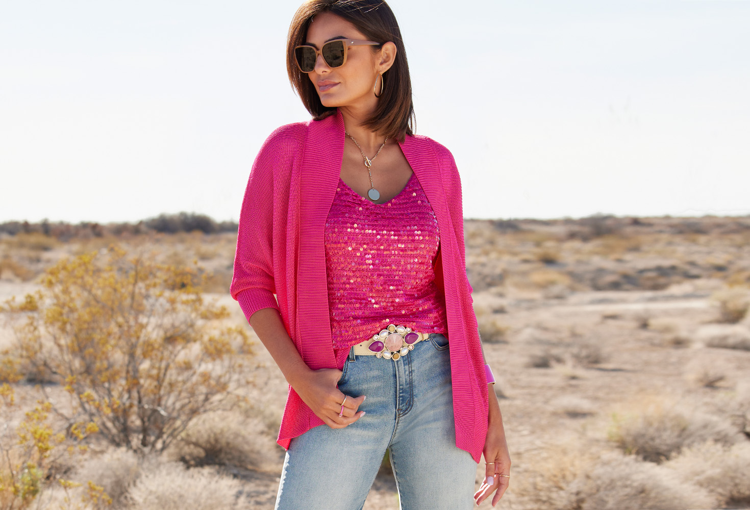 Model wearing a pink cardigan over a pink sequin embellished sweater tank, pink jewel embellished belt, and light wash jeans.