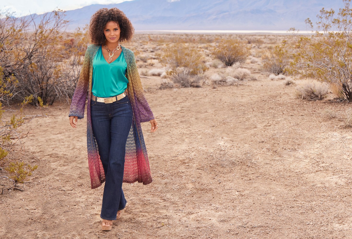 Model wearing a multicolor ombre full length duster, teal v-neck sleeveless charmeuse blouse, gold belt, dark wash straight leg jeans, and wedges.