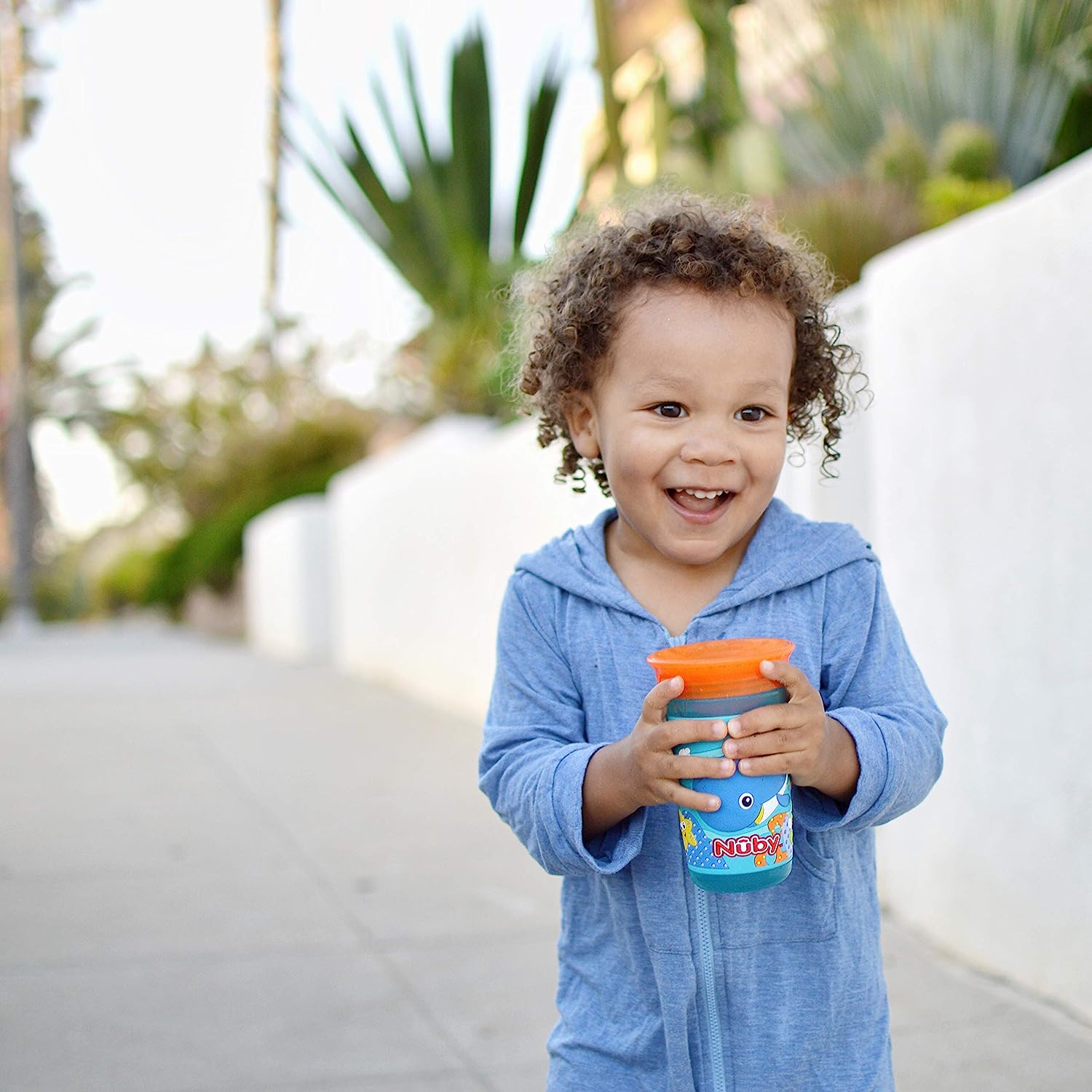 A smiling toddler standing on the sidewalk holding an orange and blue Nuby cup with a whale graphic on the outside.