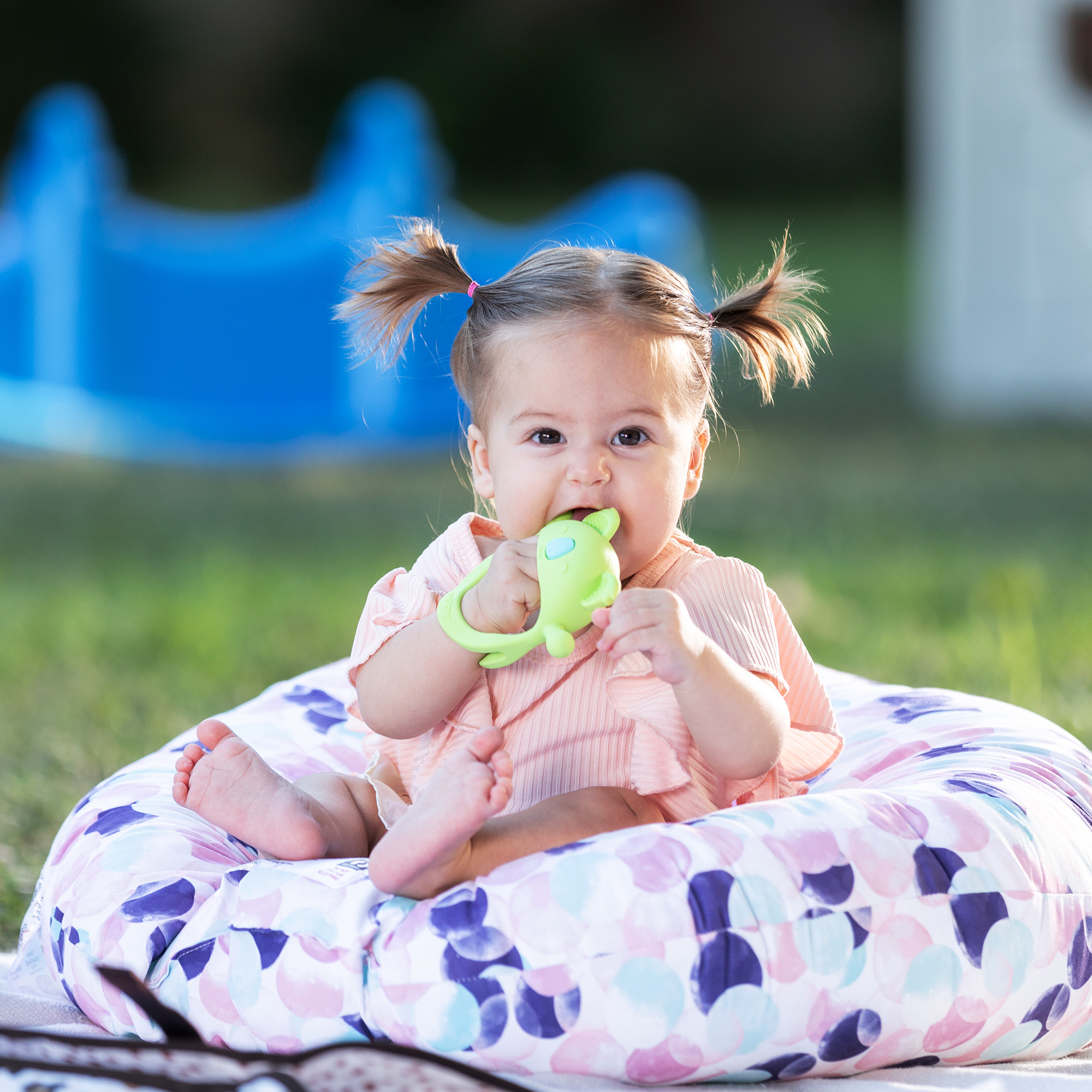 Baby girl sitting on a pillow chewing on a green koala Nuby Silicone Wrist Teething Mitten.