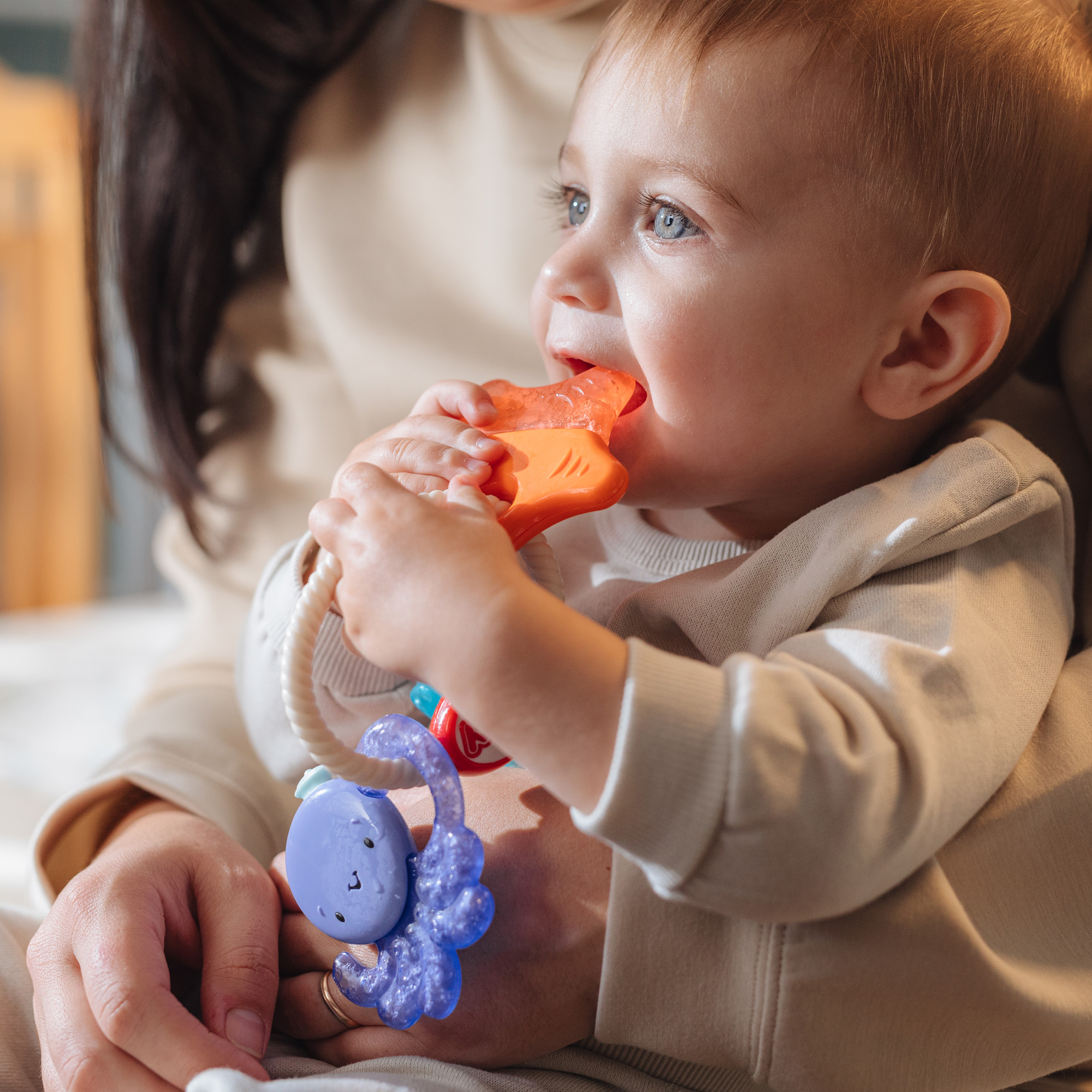 Infant using Nuby's IcyBite Sea Cooling Teether Keys while sitting on mother's lap.