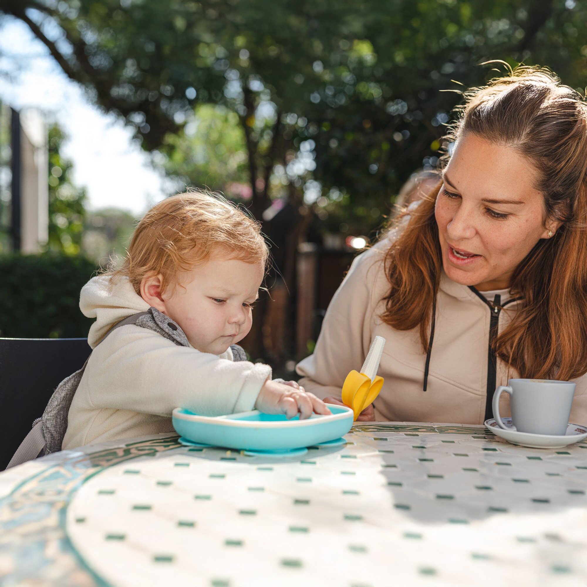 Baby eating from a Nuby Rectangular Silicone Suction Plate in aqua, holding a Silicone Banana Tube Teether, sitting next to mom.