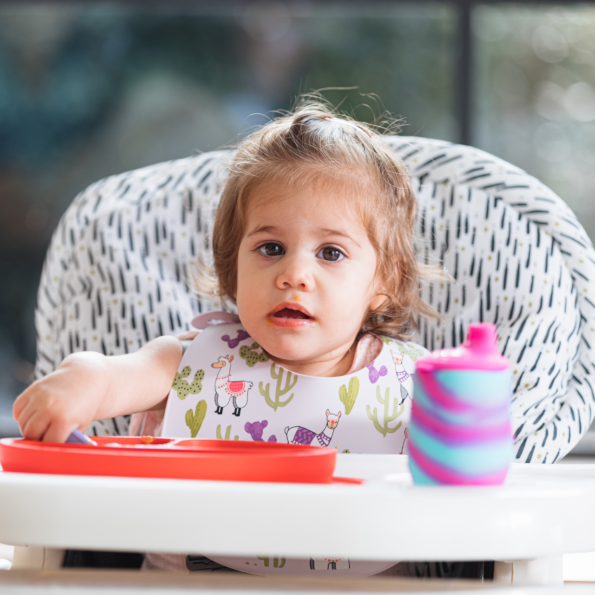 Baby sitting on Water-Resistant High Chair Cover, eating from a Red Sure Grip Miracle Mat 2-Section Plate.