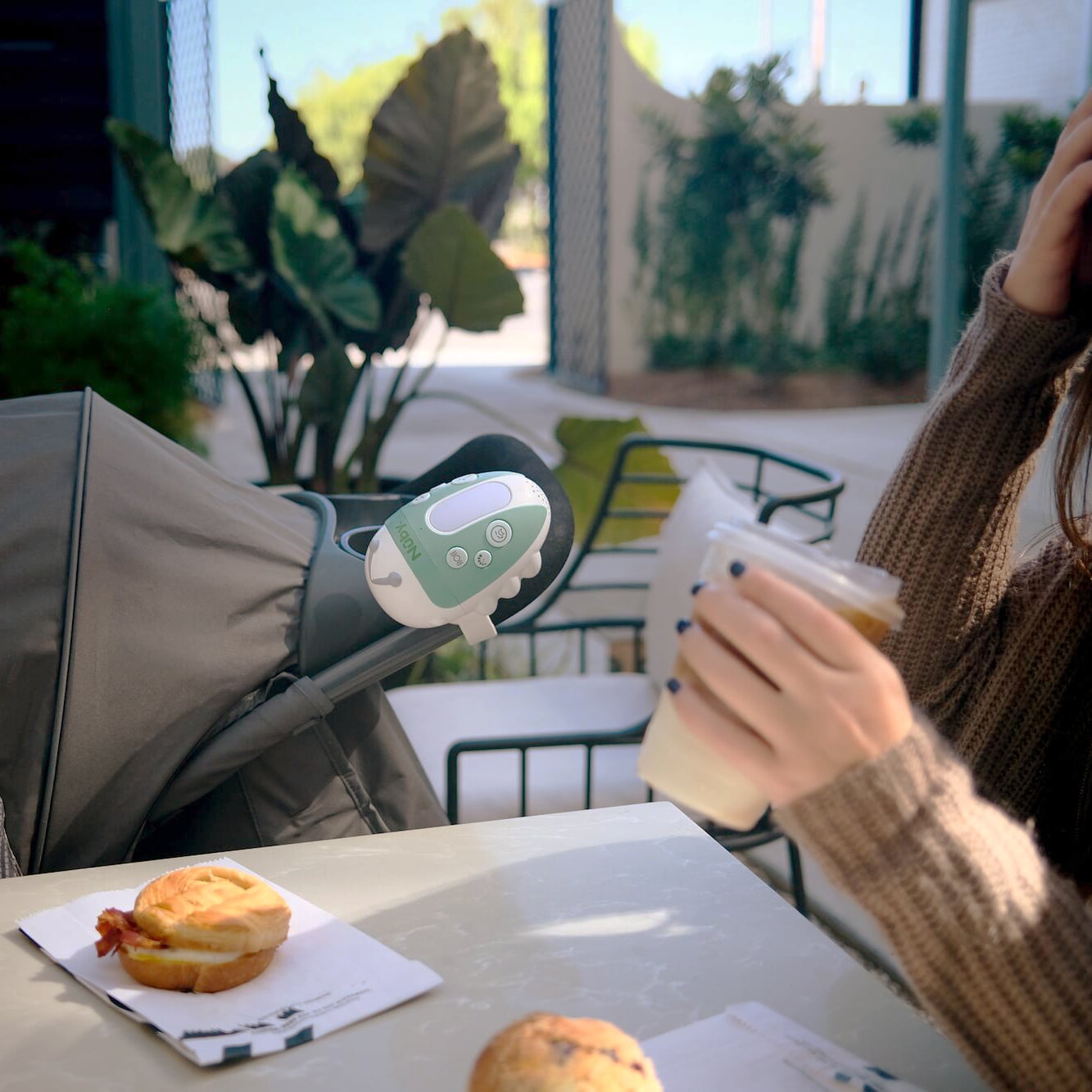 Nuby's Jitterbug stroller rocker in blue-green attached to a stroller handle, calming baby, while mom is at a café having breakfast.