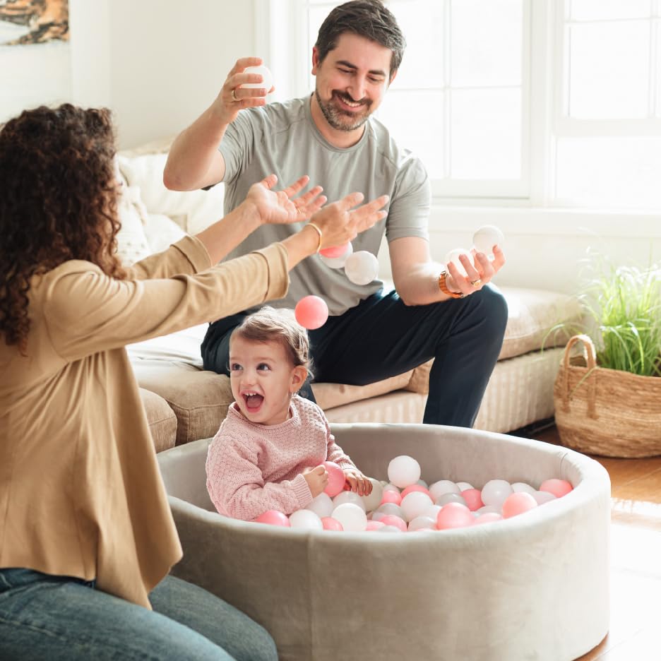 Happy toddler playing in gray Nuby ball pit with pink and white balls while smiling father and mother toss ball pit balls around.