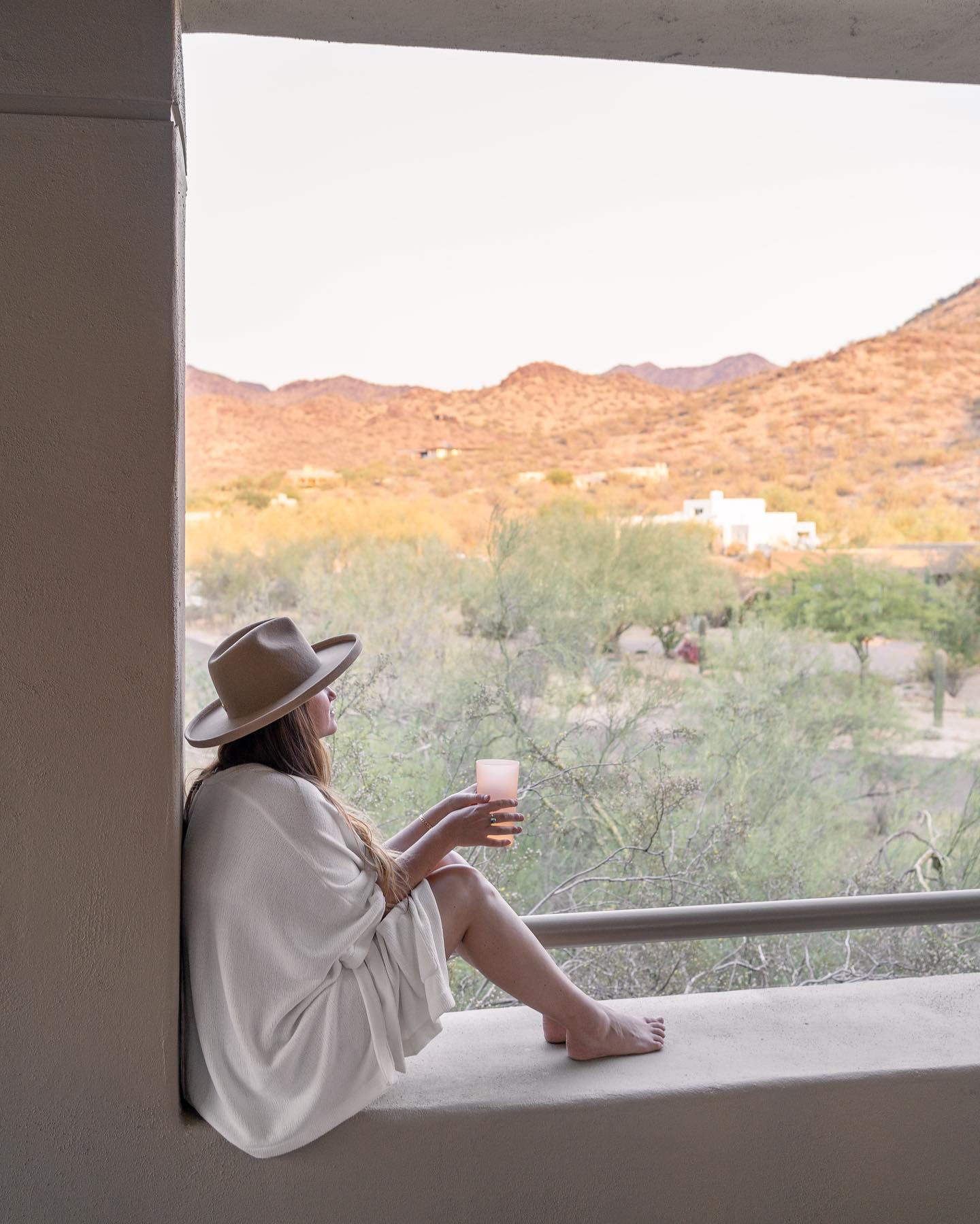 Woman looking out into desert. Woman sitting on edge of window drinking wine. Woman wearing hat and poncho. Woman wearing poncho. Woman on vacation in the desert.